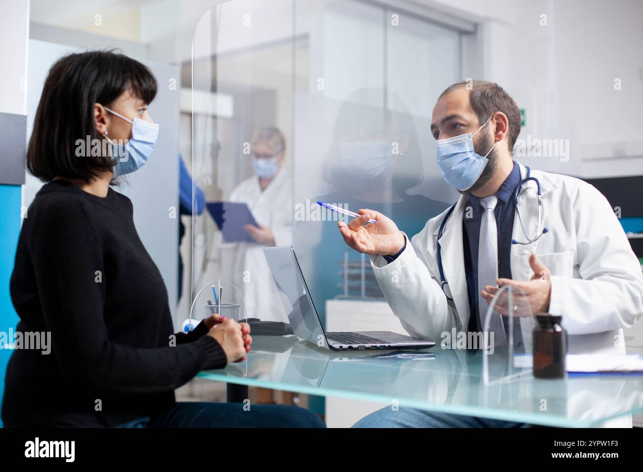 Facharzt bespricht Behandlungspläne mit der kranken Frau am Klinikpult, abgetrennt durch Kunststoffschutzschirm. Patientin, die als Ärztin in der Arztpraxis sitzt, erklärt Untersuchungsergebnisse. Stockfoto