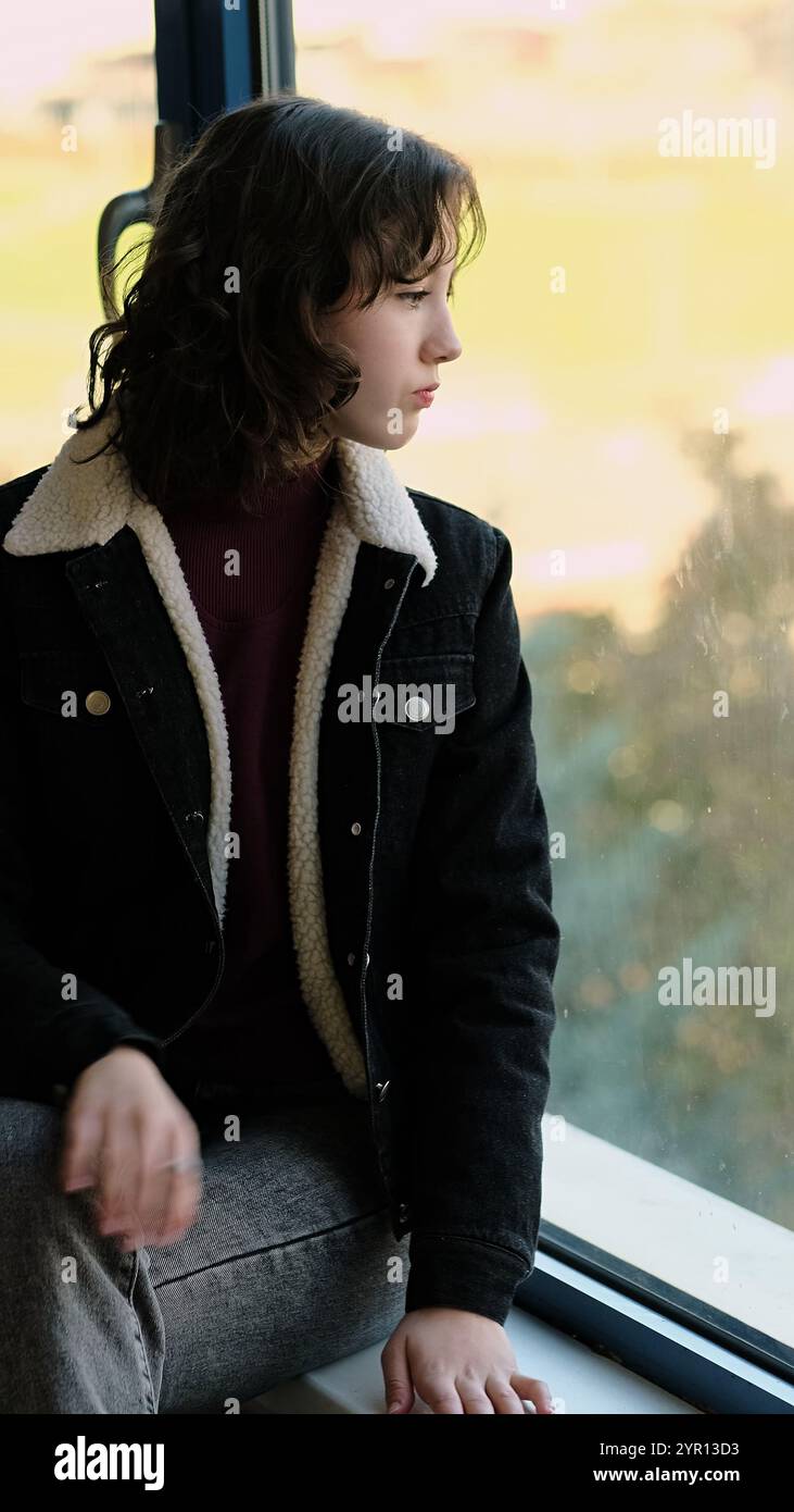 Ein ruhiges Mädchen sitzt neben einem Fenster und blickt in die Ferne mit einem reflektierenden Verhalten. Das Foto symbolisiert emotionale Tiefe, Verbindung zur Natur Stockfoto