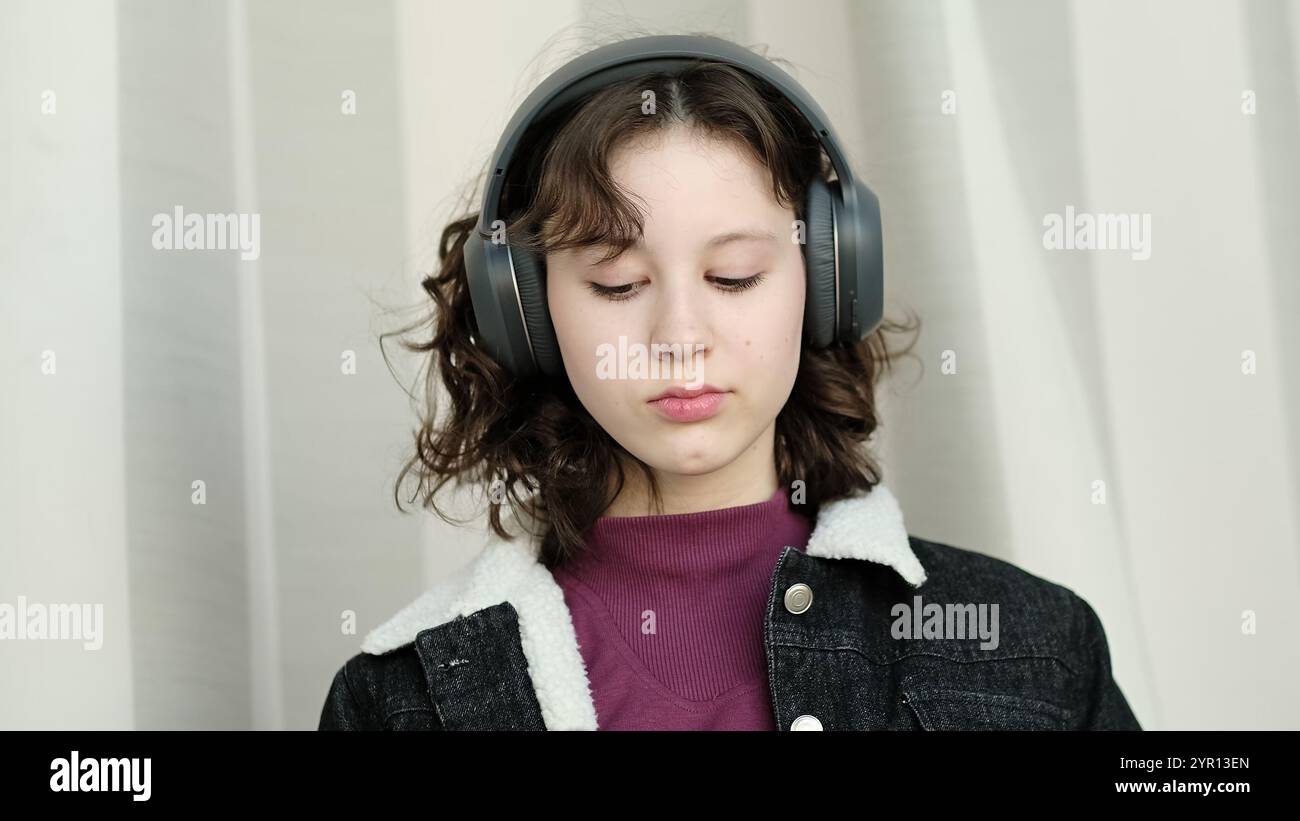 Ein nachdenkliches Mädchen mit Kopfhörern entspannt sich in einem lichtdurchfluteten Raum, ihr Ausdruck ist ruhig. Das Bild unterstreicht den persönlichen Musikgenuss und Stockfoto