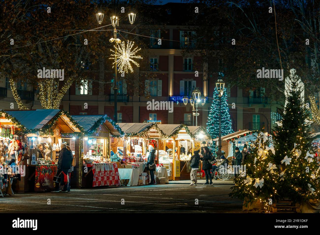 Leute kaufen auf einem traditionellen festlichen Weihnachtsmarkt mit Holzbuden mit Weihnachtsbeleuchtung und Dekoration am Abend in Asti, Italien. Stockfoto