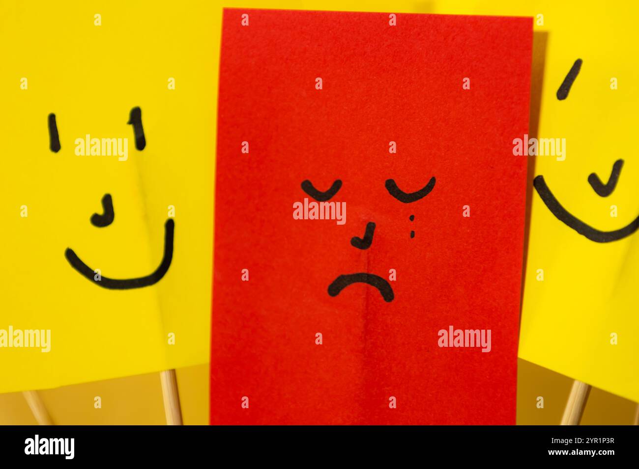 Nahaufnahme von rotem Papier mit traurigem depressivem Gesicht, umgeben von gelben Smiley-Papieren, Konzept von Depression und Isolation von der Gesellschaft Stockfoto