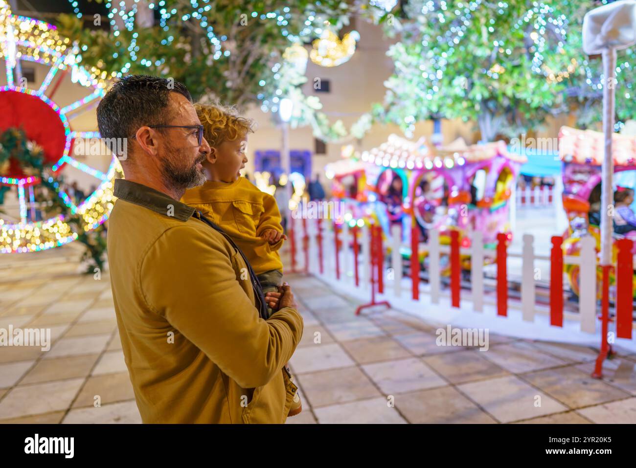 Vater hält seinen Sohn in der Hand und beobachtet eine Karussellfahrt auf einem weihnachtsmarkt und genießt die festliche Atmosphäre Stockfoto