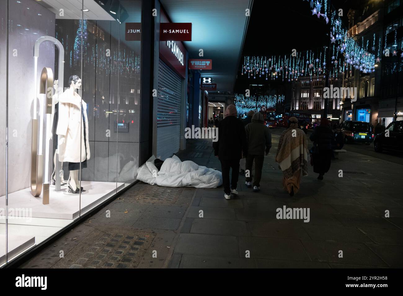 Fußgänger passieren einen Obdachlosen, der in einer Bettdecke vor einem Schaufenster in der Oxford Street im Zentrum von London schläft Stockfoto