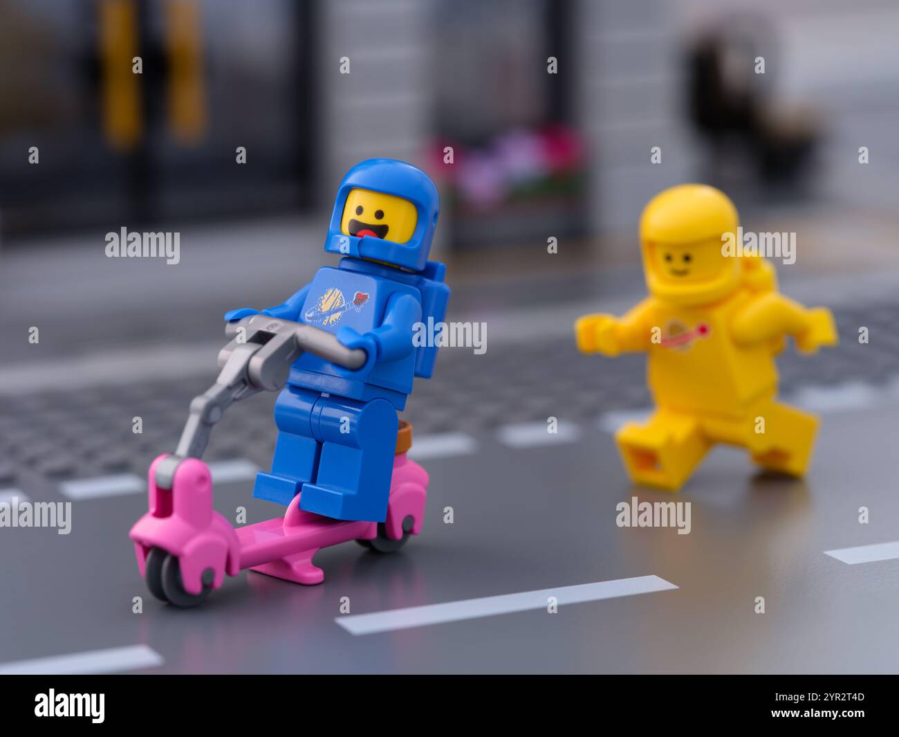 Tambow, Russische Föderation - 12. Juli 2024 gelbe Lego-Astronautenfigur folgt einem blauen Astronauten, der auf einer Straße mit einem Roller fährt Stockfoto