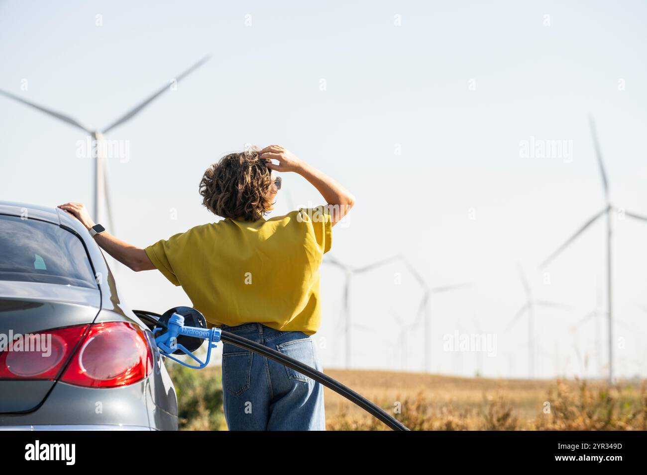 Eine Frau mit einem Wasserstoff-Auto. Betanken des Autos mit Wasserstoffkraftstoff. Windräder im Hintergrund. Stockfoto