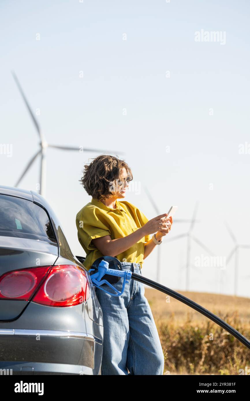 Eine Frau mit einem Wasserstoff-Auto. Betanken des Autos mit Wasserstoffkraftstoff. Windräder im Hintergrund. Stockfoto