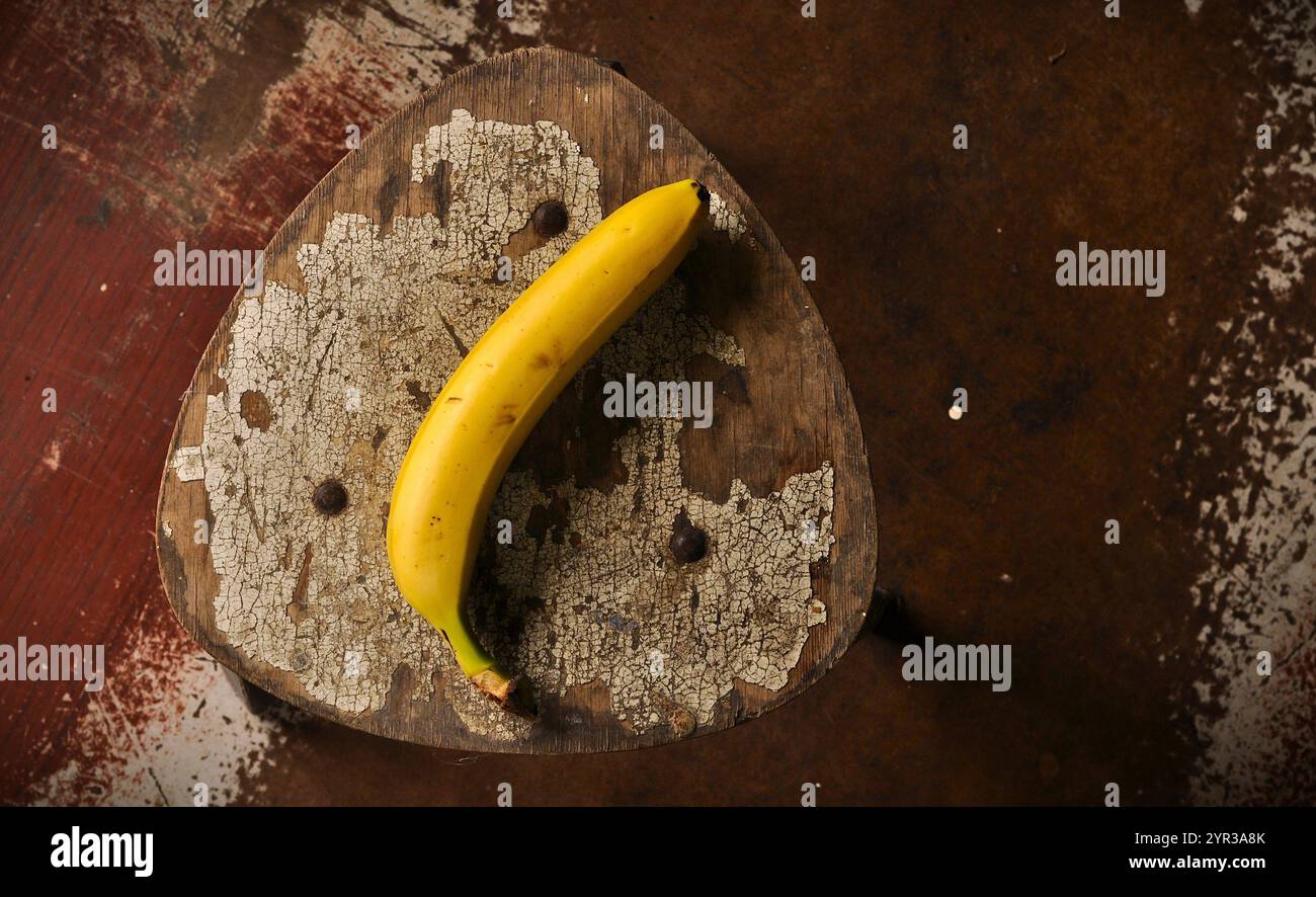 Schäbiger Stuhl und Banane. Bizarre Tapete. Alter, abgenutzter Holzstuhl mit abblätternder Farbe und rostigen Details. Dunkler, dramatischer Hintergrund. Künstlerische Tapete Stockfoto