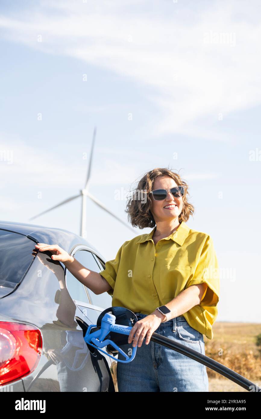 Eine Frau mit einem Wasserstoff-Auto. Betanken des Autos mit Wasserstoffkraftstoff. Windräder im Hintergrund. Stockfoto