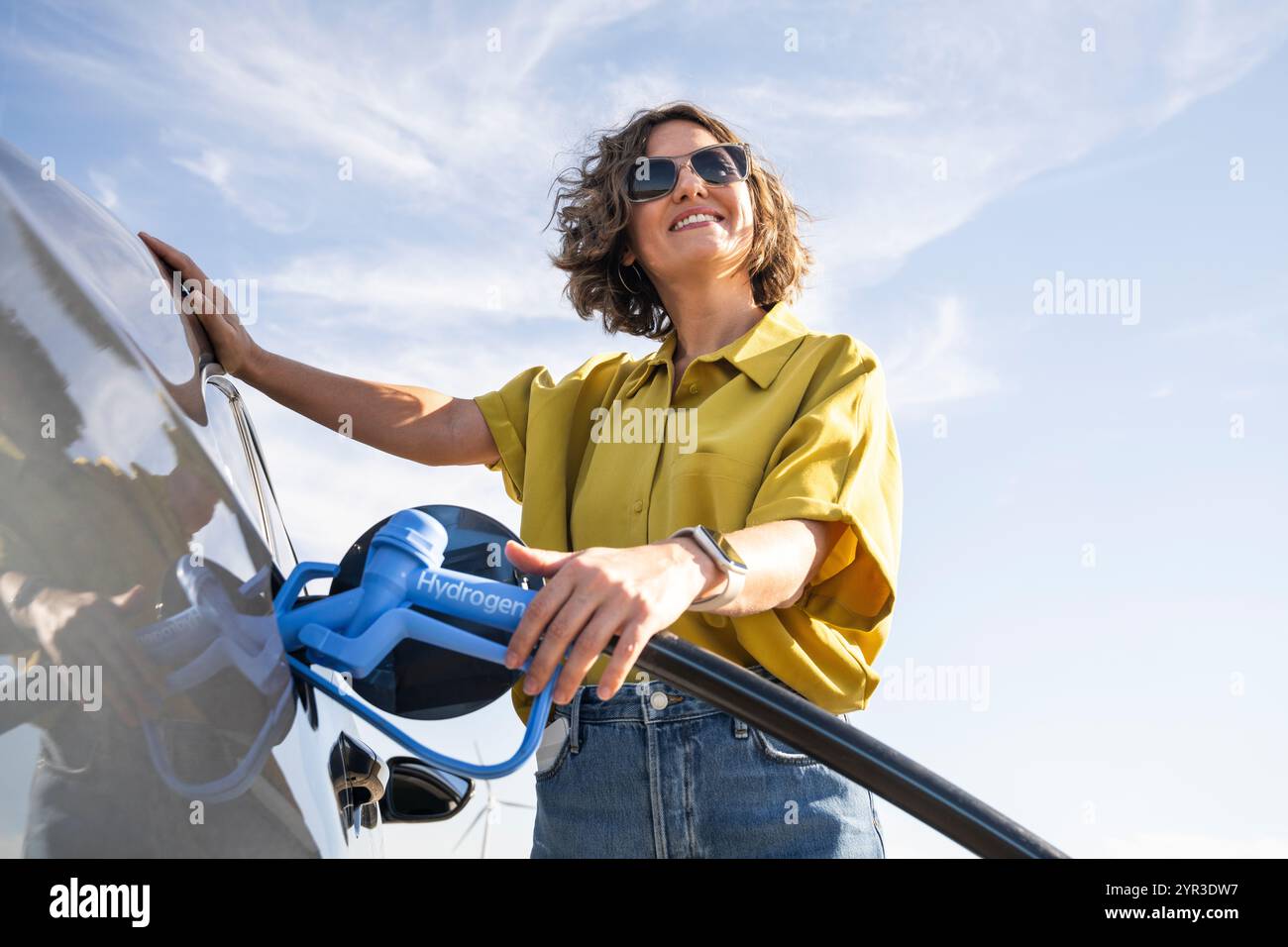 Eine Frau mit einem Wasserstoff-Auto. Betanken des Autos mit Wasserstoffkraftstoff. Windräder im Hintergrund. Stockfoto