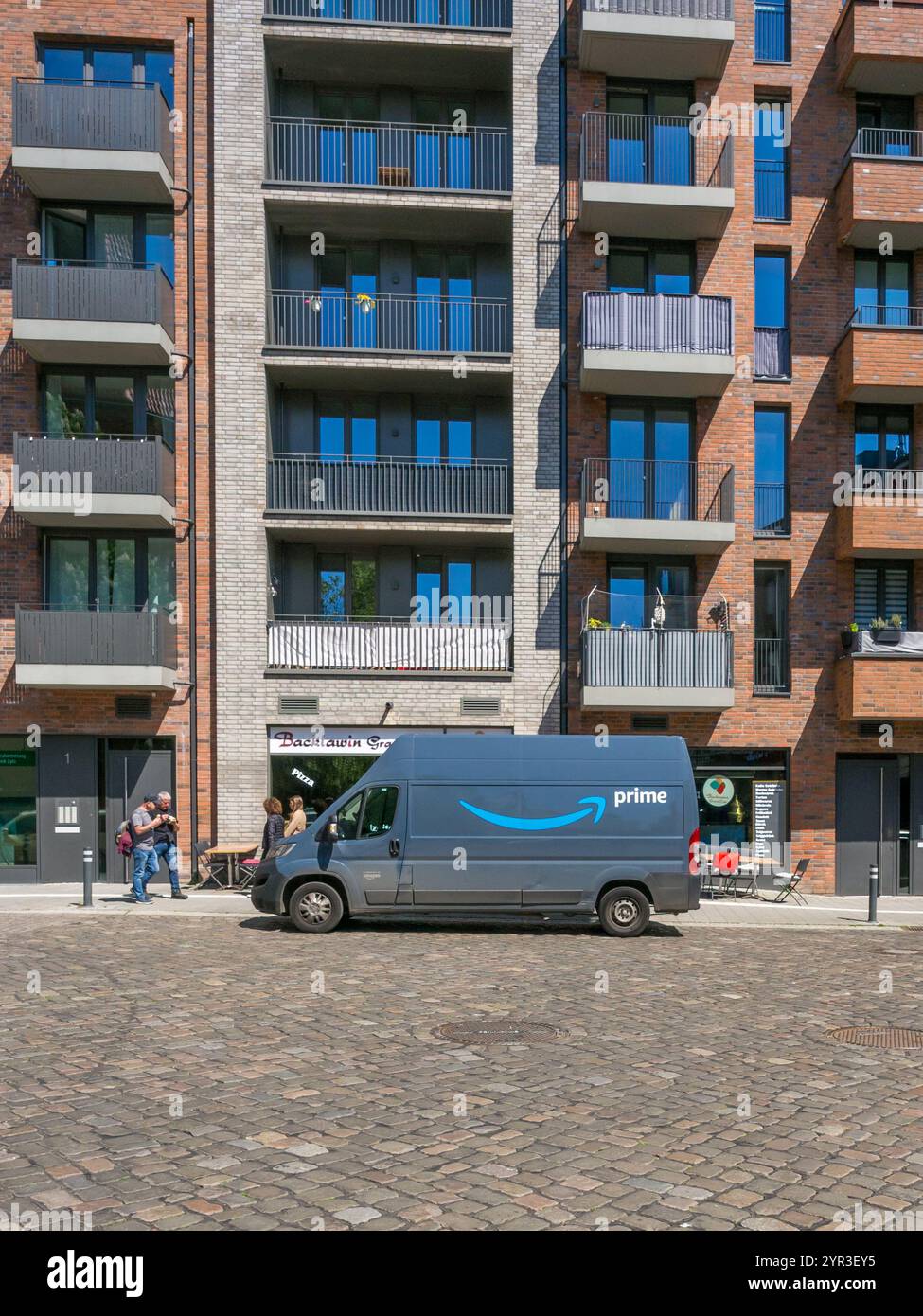 Ein Amazon Prime Lieferwagen parkte auf einer Kopfsteinpflasterstraße vor einem Geschäft, über dem an einem hellen, sonnigen Tag ein Wohnhaus mit Balkonen steht. Stockfoto