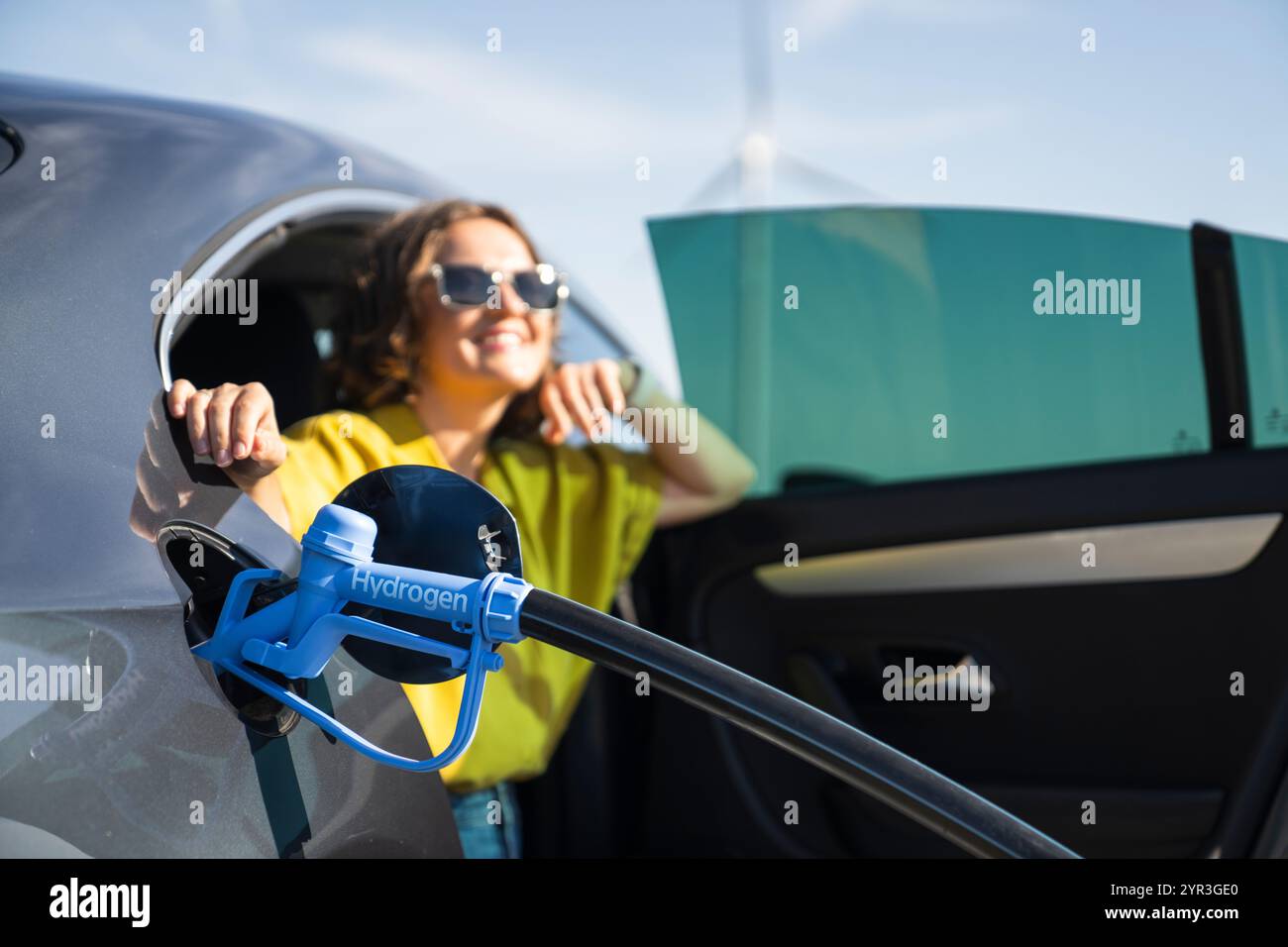 Eine Frau mit einem Wasserstoff-Auto. Betanken des Autos mit Wasserstoffkraftstoff. Windräder im Hintergrund. Stockfoto