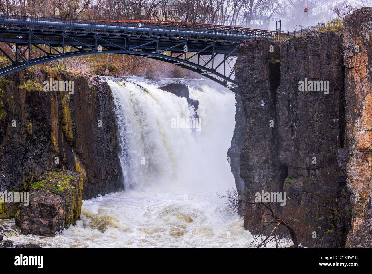 Der Wasserfall Great Falls in Paterson, New Jersey, stürzt mächtig über felsige Klippen mit einer eisernen Brücke darüber. Stockfoto