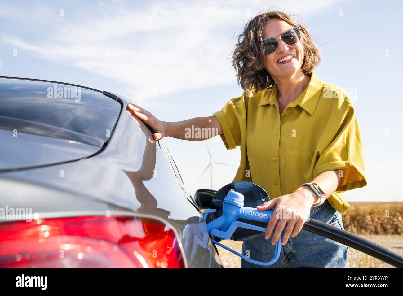 Eine Frau mit einem Wasserstoff-Auto. Betanken des Autos mit Wasserstoffkraftstoff. Windräder im Hintergrund. Stockfoto