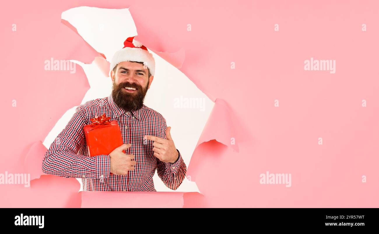 Weihnachten oder Neujahr einkaufen. Lächelnder bärtiger Mann mit Weihnachtsmütze und kariertem Hemd, der mit dem Finger auf das Weihnachtsgeschenk zeigt und durch ein Loch in Papier schaut. Chri Stockfoto