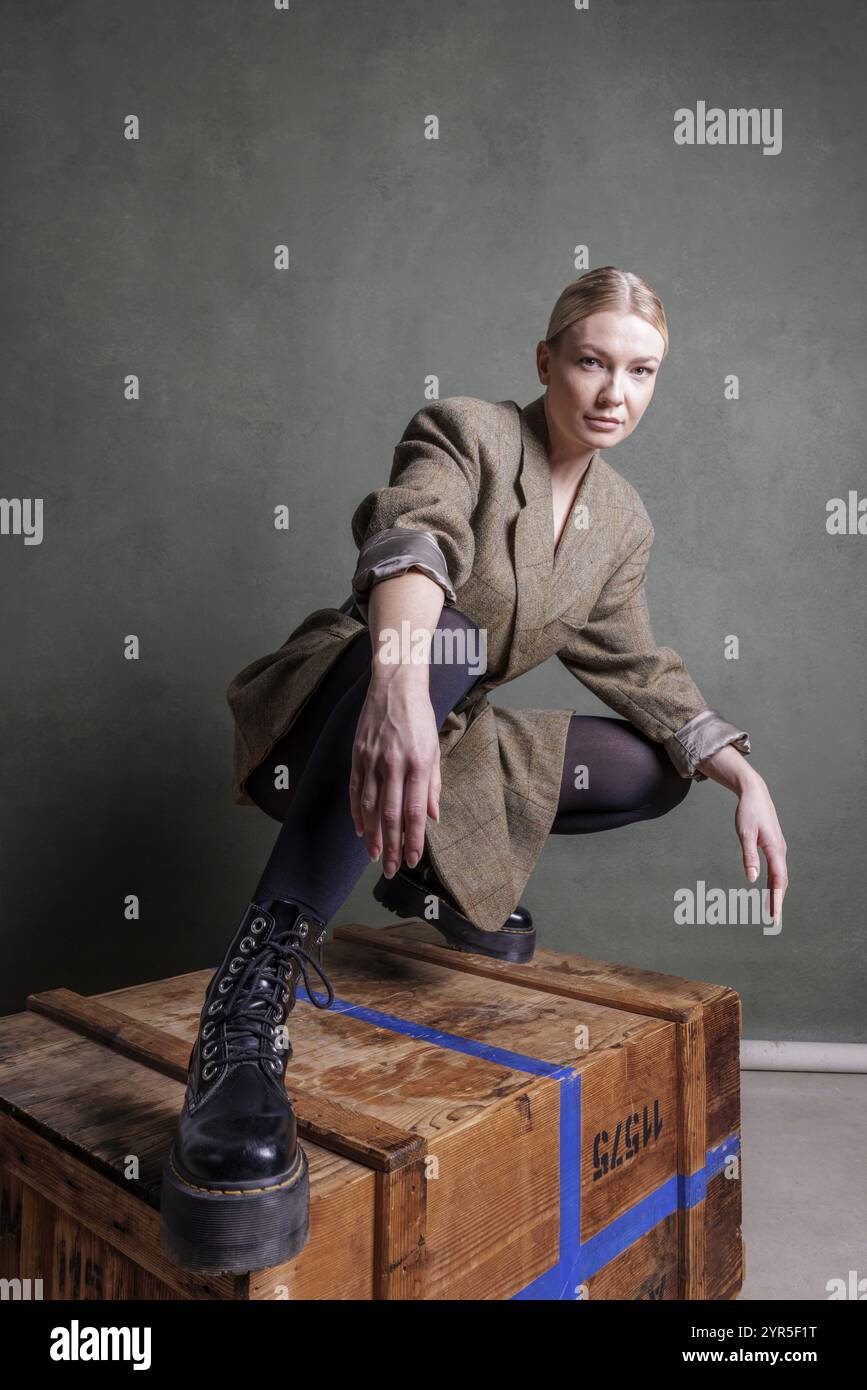 Frau posiert selbstbewusst auf einer Holzkiste, elegant gekleidet vor einem grünen Hintergrund, Fotoshooting Stockfoto