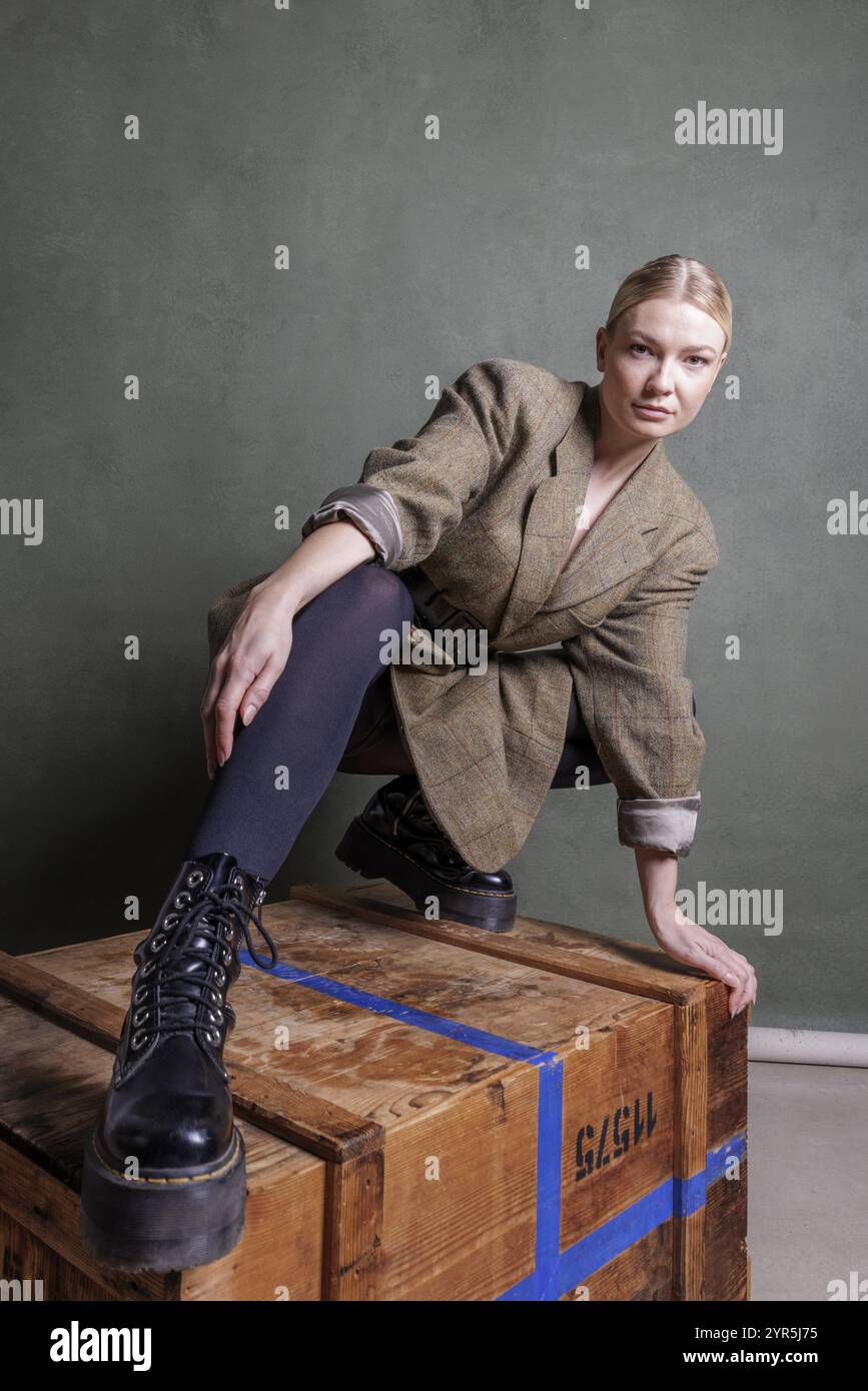 Eine stilvoll gekleidete Frau posiert auf einer Holzkiste vor einem grünen Hintergrund, Fotoshooting Stockfoto