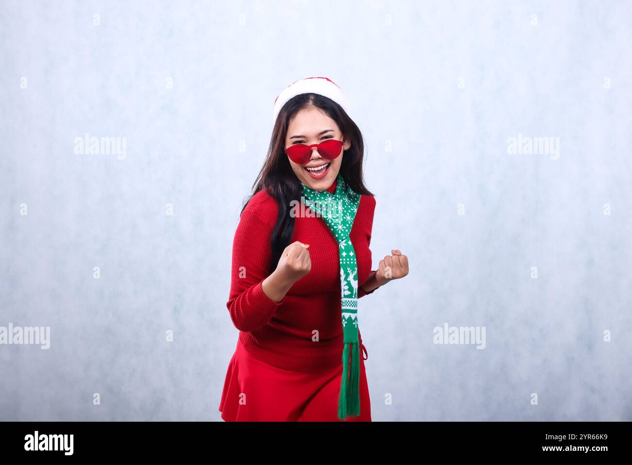 Charmante asiatische Frau mit rotem Pullover mit Weihnachtsmütze und Schal, fröhlicher Geist zur Kamera mit Brille, beide Hände nach vorne geballt, feiern Stockfoto