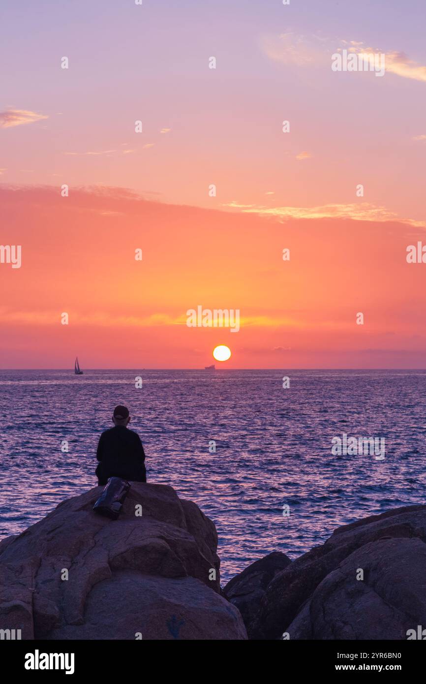 Der junge Mann sitzt auf einem Felsen am Meer und genießt einen atemberaubenden Sonnenuntergang über einem ruhigen Meer, mit weit entfernten Segelbooten, die die ruhige Atmosphäre verstärken Stockfoto