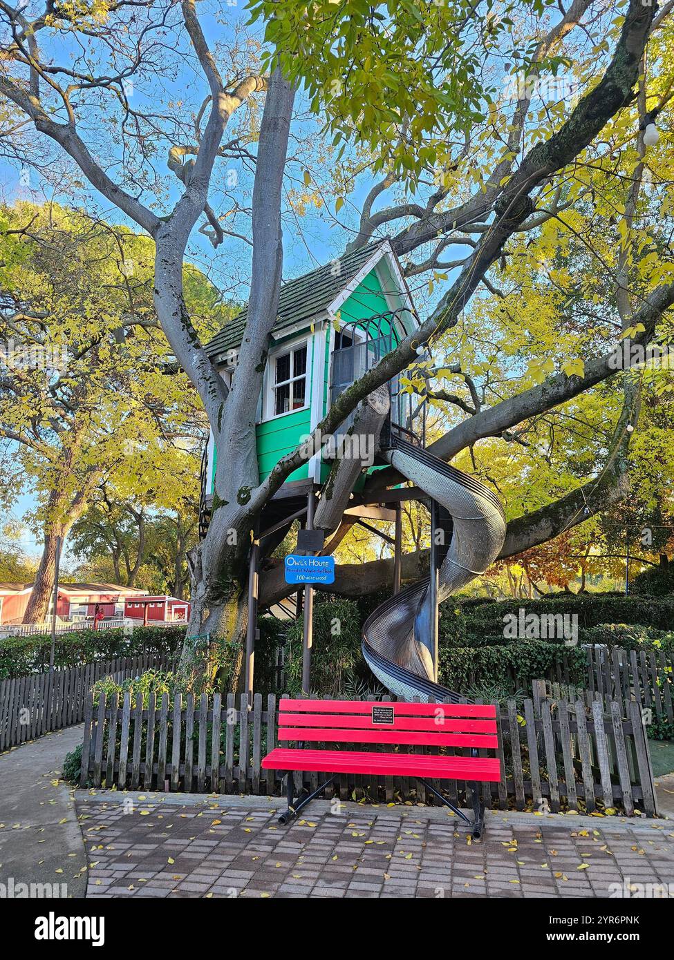 Bezauberndes Baumhaus in einem großen Baum mit einer spiralförmigen Rutsche und einer hellroten Bank in einer friedlichen Parklandschaft. Perfekt für Themen der Kindheit. Stockfoto Bezauberndes Baumhaus in einem großen Baum mit einer spiralförmigen Rutsche und einer hellroten Bank in einer friedlichen Parklandschaft. Perfekt für Themen der Kindheit. Stockfoto