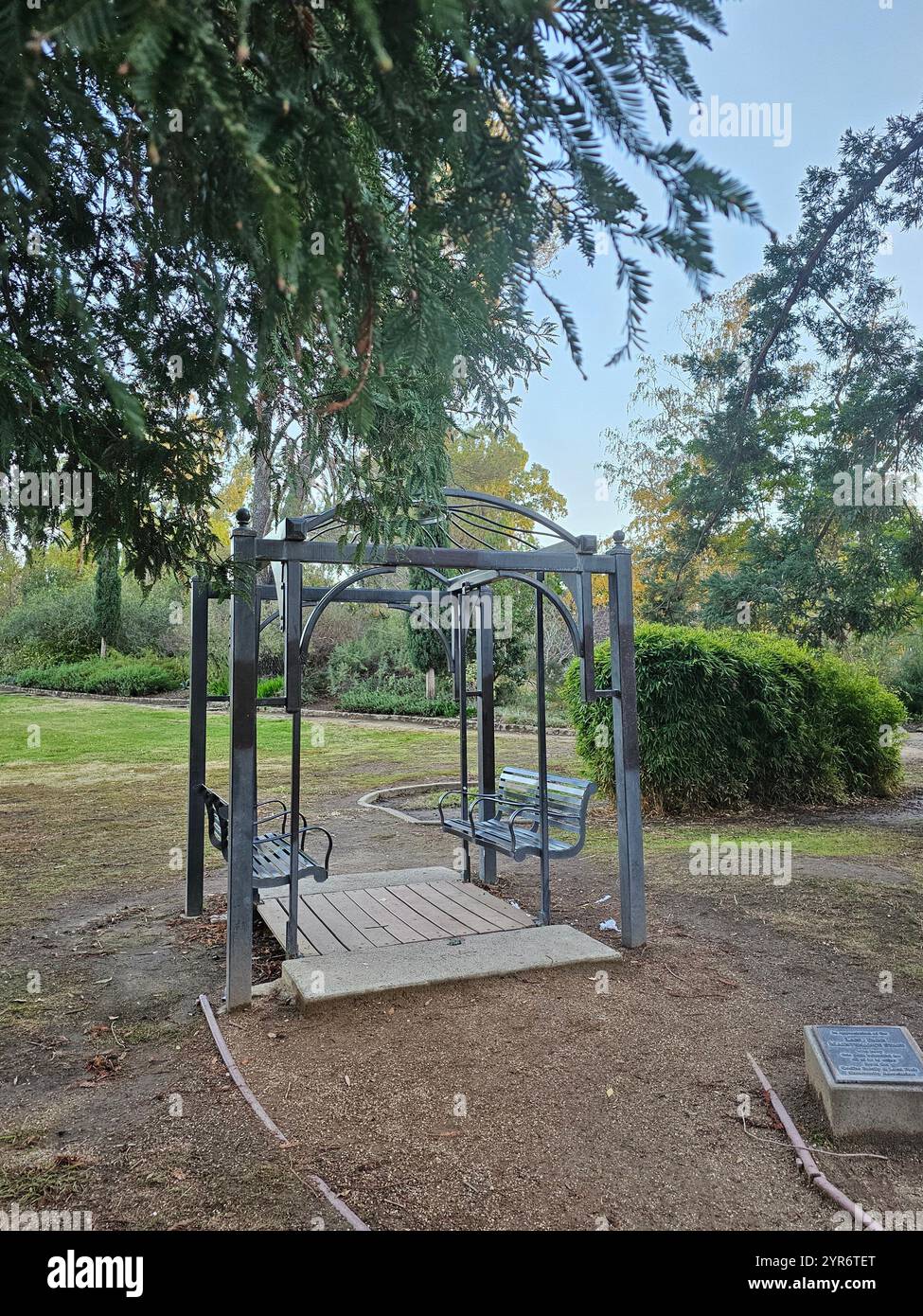 Metallschaukel unter einer Pergola in einer ruhigen Parklandschaft, umgeben von Grün und Bäumen. Ein ruhiger Ort für Entspannung und Outdoor-Genuss. - Smartphone-aufgenommenes Stockfoto