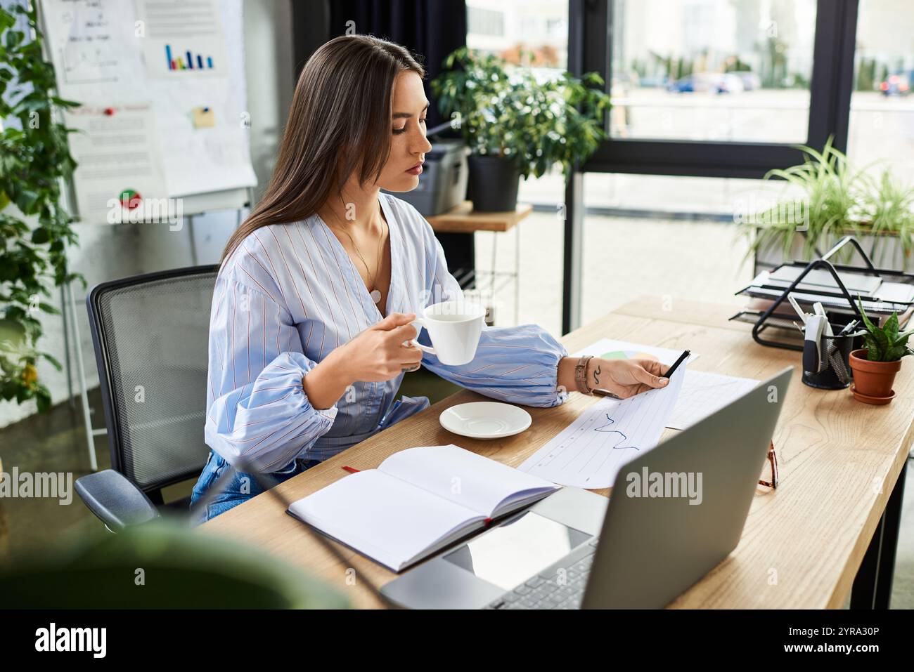 Brünette arbeitet intensiv in einem hellen, mit Pflanzen gefüllten Büro mit Kaffee in der Hand. Stockfoto