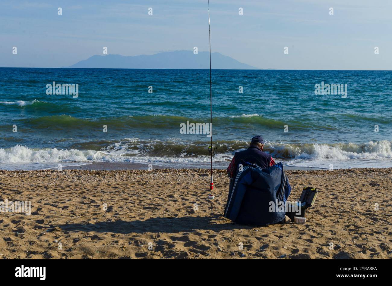 Mann im Ruhestand, der an einem Sandstrand mit Meer und Insel im Hintergrund sitzt Stockfoto