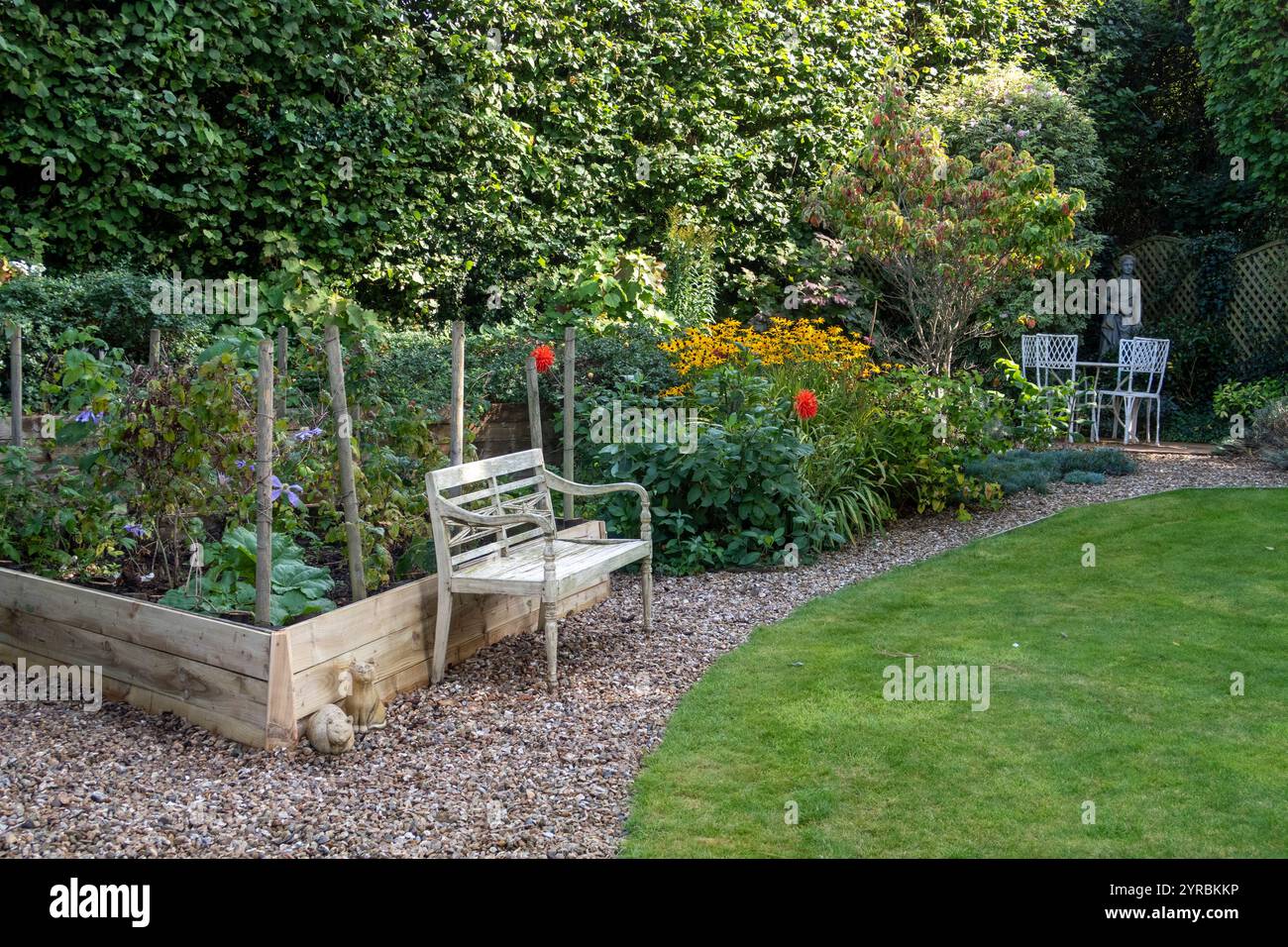 Gartenszene mit erhöhtem Obstbeet, Kieswegen, heißem Garten - Rudbeckia und Dahlien, Möbeln, Figuren, Rasen. Stockfoto