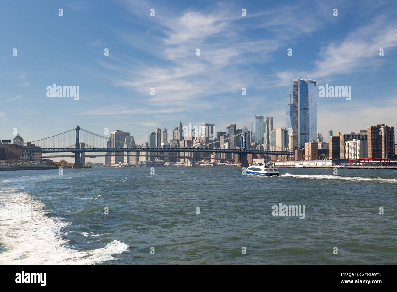 Erleben Sie die Skyline von New York City von der Fähre aus an einem sonnigen Tag. Majestätisch erheben sich Wolkenkratzer, die Manhattan Bridge überquert das Wasser Stockfoto