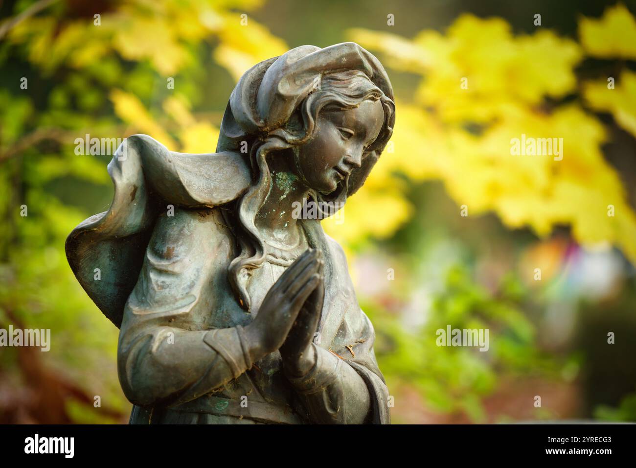 Liebevoll und liebevoll aussehende Statue einer Frau mit betenden Händen auf einem Grabstein eines Friedhofs vor glühenden Herbstblättern in einem verschwommenen Hintergrund Stockfoto