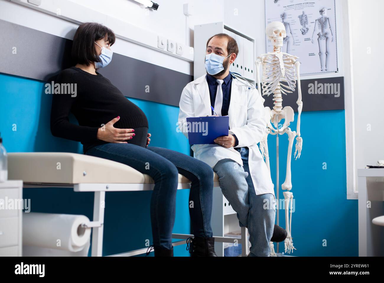 Junger Gesundheitsspezialist hält Klemmbrett und berät schwangere Patientin und bespricht Diagnosedetails in der Klinik. Der männliche Arzt sitzt neben der werdenden Mutter auf dem Krankenhausbett und bietet eine Mutterschaftsberatung an. Stockfoto