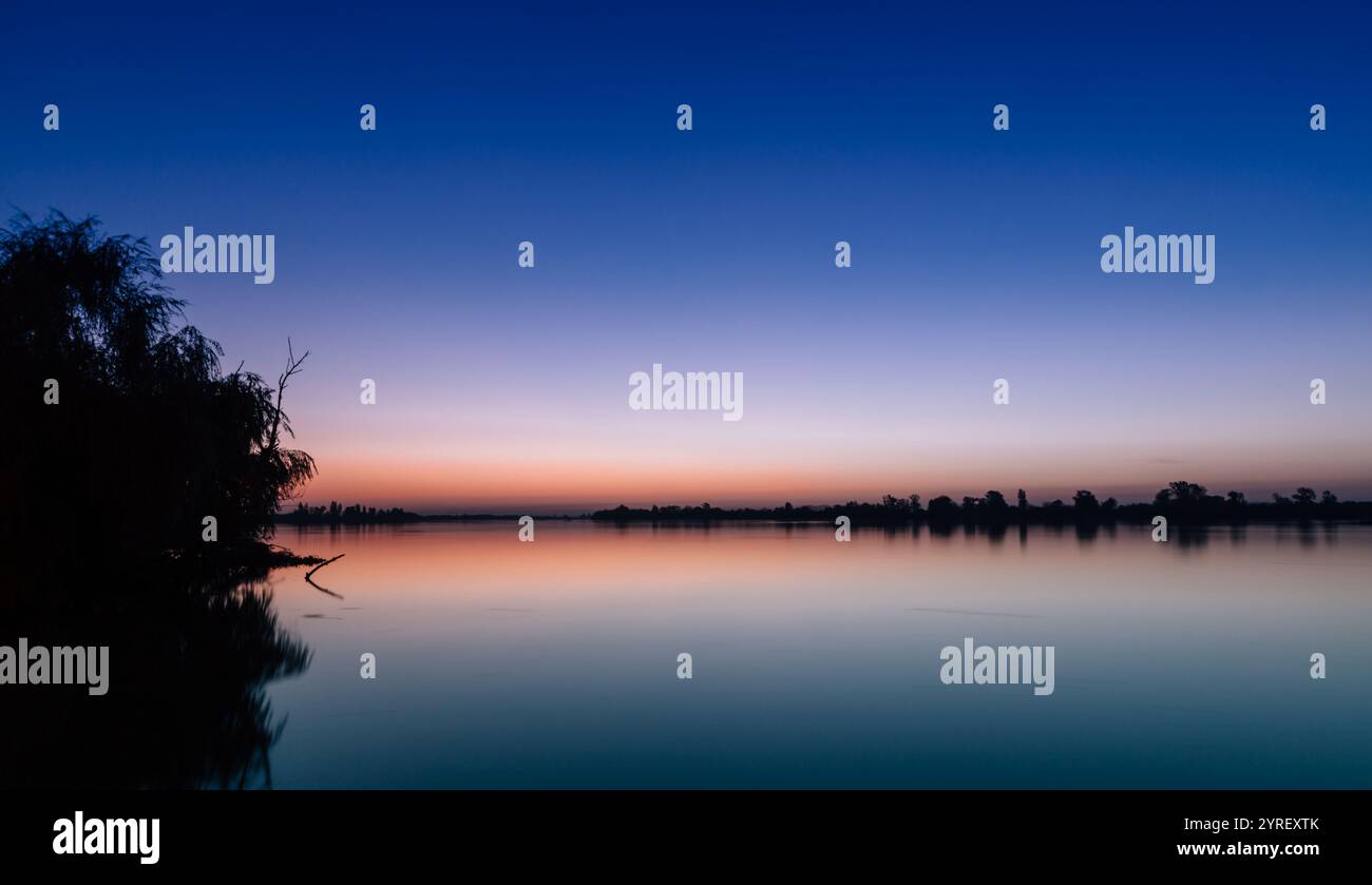 Die leuchtenden Farben des Sonnenuntergangs spiegeln sich auf dem ruhigen See wider und schaffen eine friedliche Szene mit Baumsilhouetten, die Ruhe verleihen Stockfoto