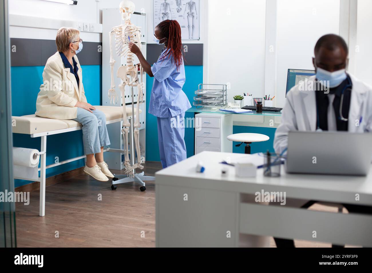 afroamerikanische Krankenschwester zeigt auf Skelett, das älteren Patienten die Gesundheit der Wirbelsäule erklärt. Weibliche medizinische Assistentin in Gesichtsmaske beschreibt die Anatomie der alten weißen Frau anhand des Skelettmodells als Referenz. Stockfoto