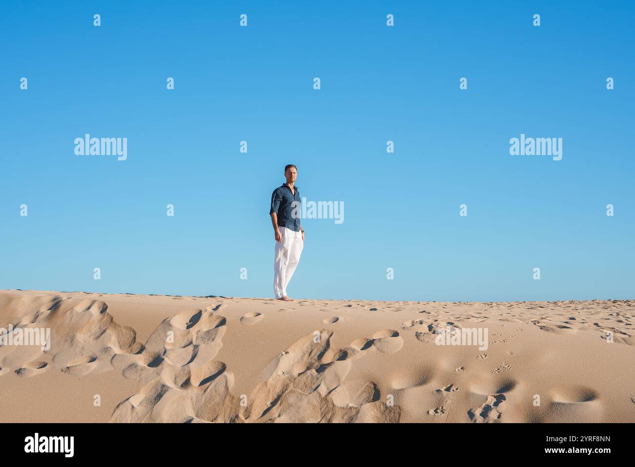 Ein Mann in dunklem Hemd und weißer Hose steht auf einer Sanddüne in Tarifa, Spanien. Die Szene bietet glatten Sand und einen hellen, wolkenlosen Himmel, der einen erzeugt Stockfoto