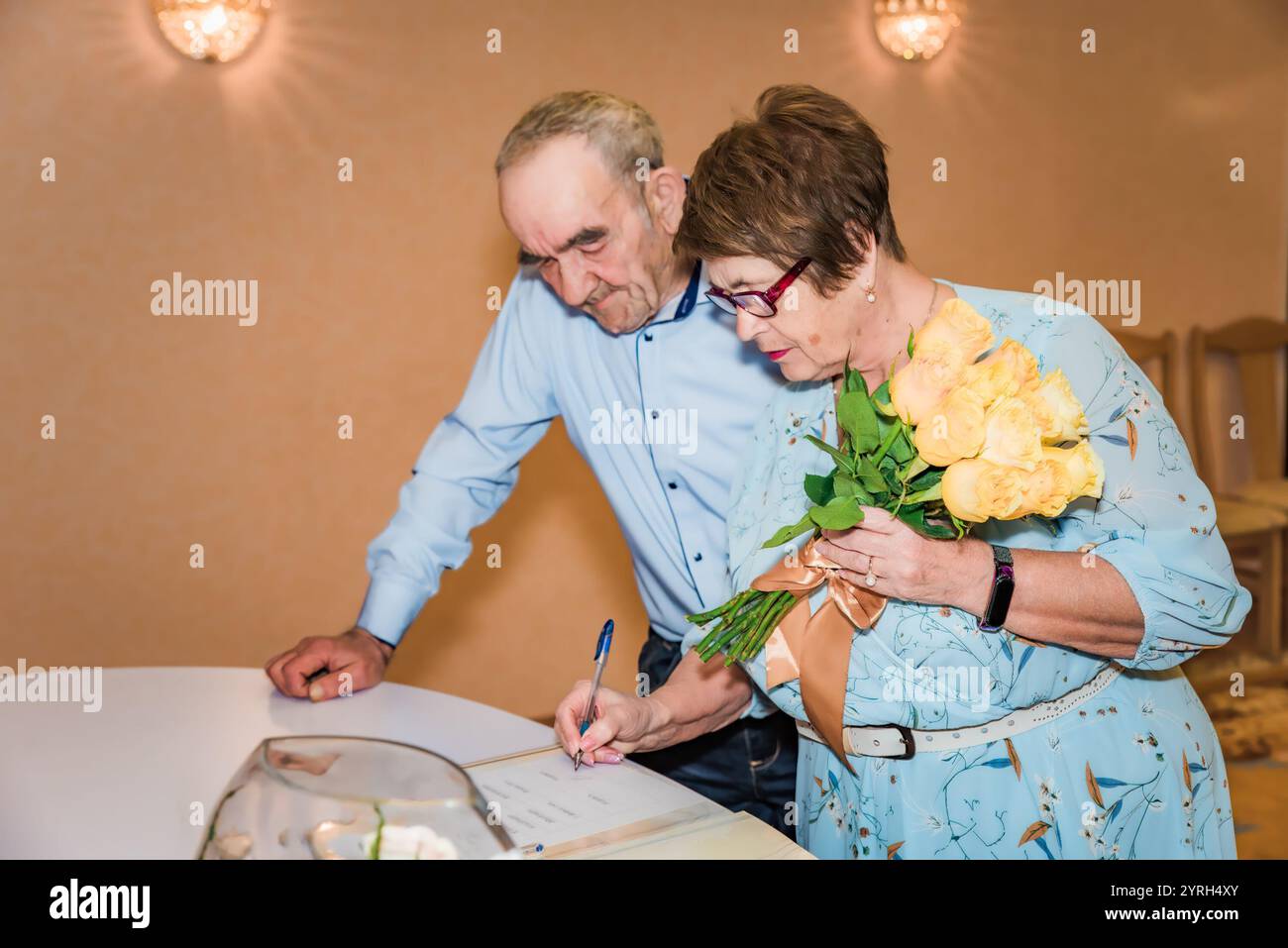 Ein älteres Ehepaar unterschreibt Dokumente in einem Standesamt in Russland, um ihren 50. Hochzeitstag zu feiern. Die Frau hält einen Blumenstrauß aus gelber Rose Stockfoto