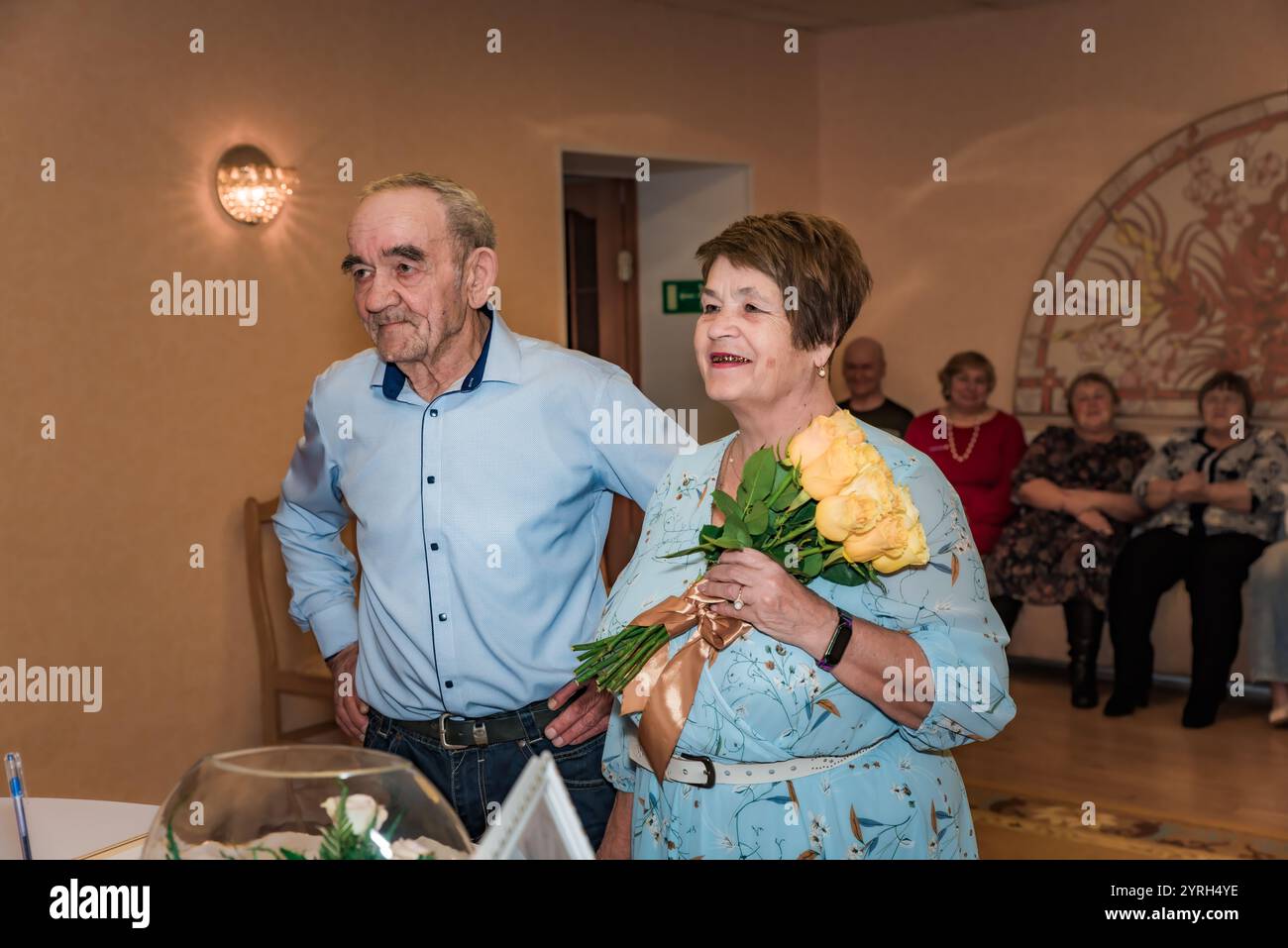 Ein Paar feiert seinen 50. Hochzeitstag (goldene Hochzeit) im Standesamt. Die Frau lächelt, während sie einen Blumenstrauß hält, und ihre Freunde Stockfoto