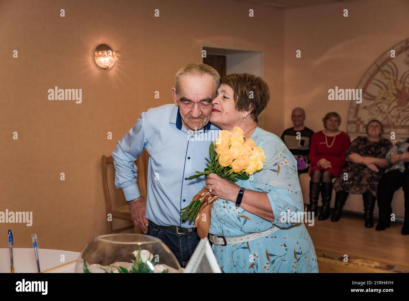 Das Paar feiert seinen 50. Hochzeitstag (goldene Hochzeit) im Standesamt. Die Frau küsst ihren Mann auf die Wange, während sie einen bou hält Stockfoto