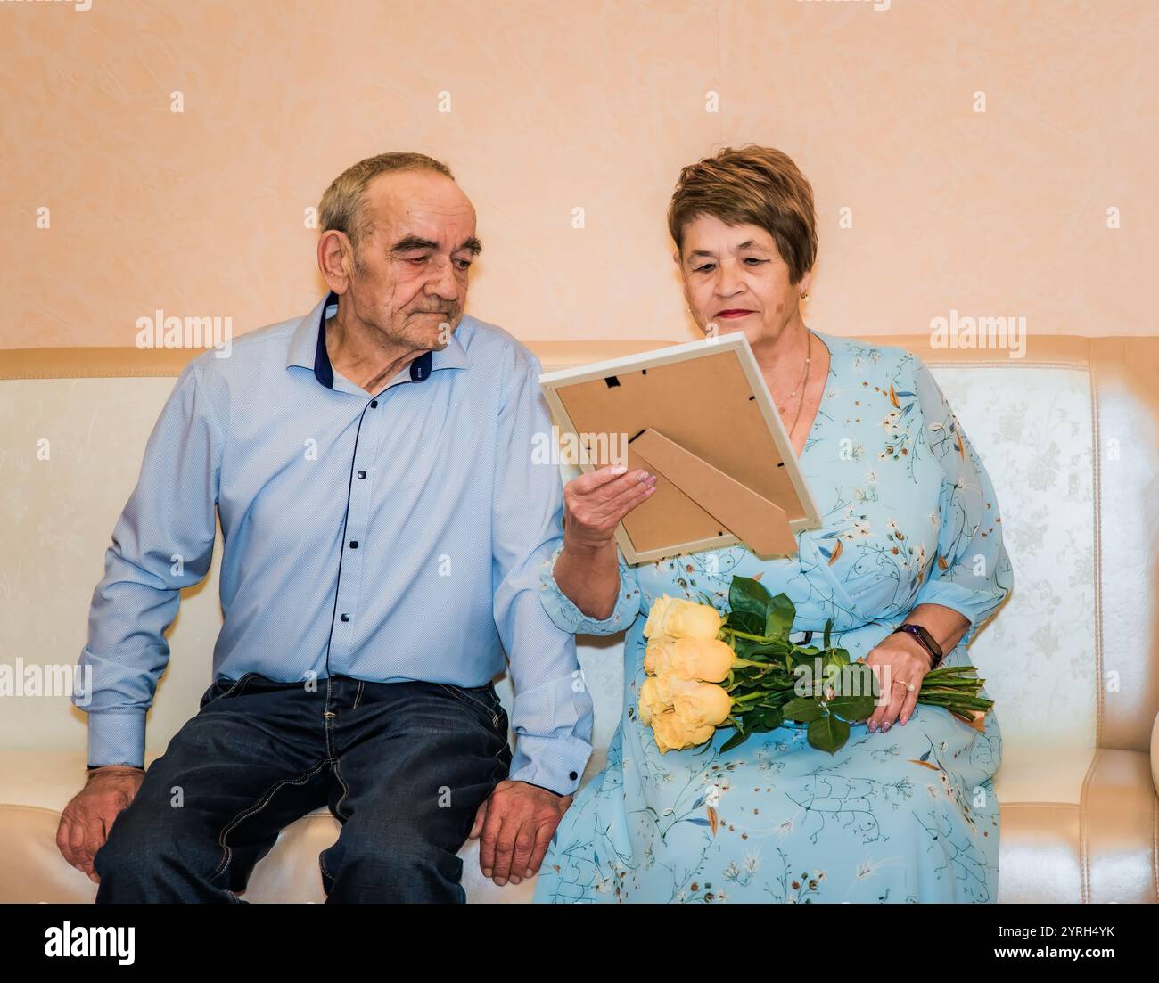 76-jähriger Mann und 71-jährige Frau feiern ihren 50. Hochzeitstag. Die Frau in einem blauen Kleid hält gelbe Rosen und sie sehen sich ein Geschenk an Stockfoto