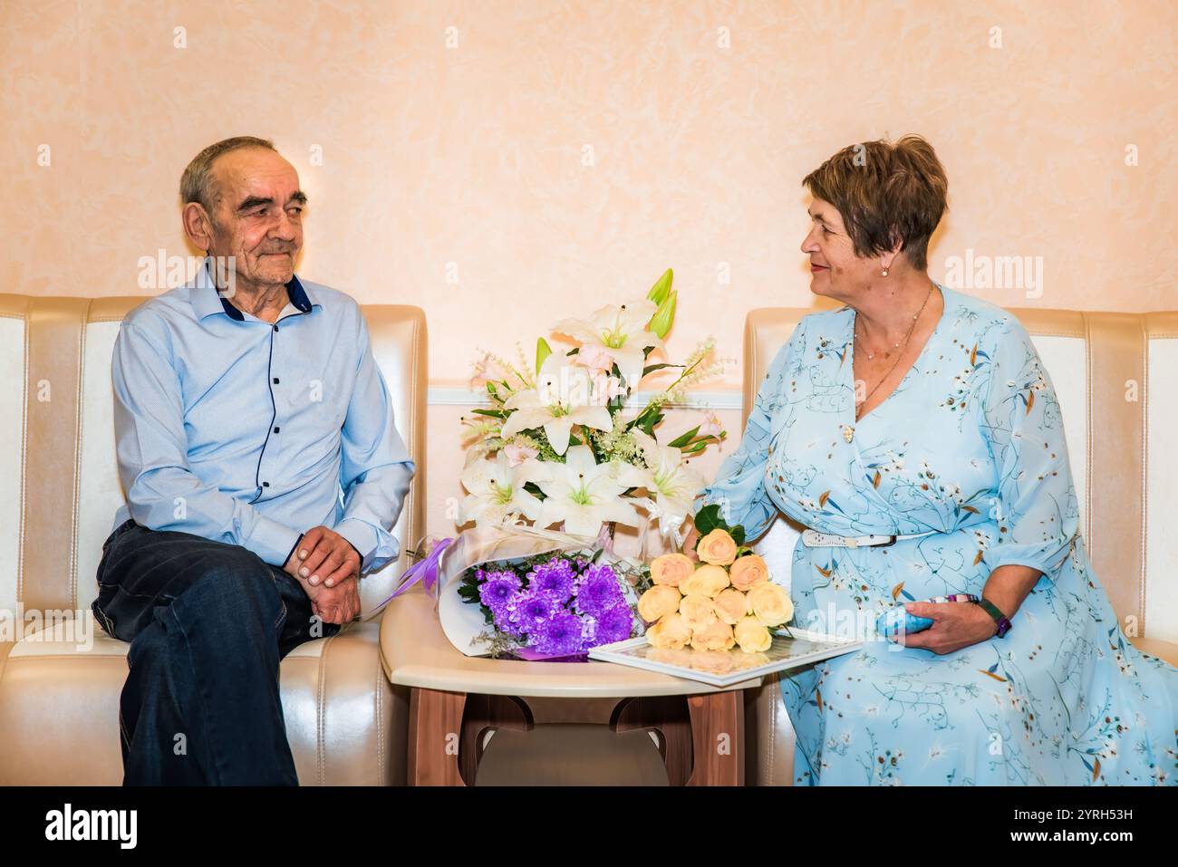 Ein russisches Paar über 70, das ihren goldenen Hochzeitstag im Standesamt feiert, sitzt zusammen auf Sofas. Sie lächeln einander an, umgeben Stockfoto