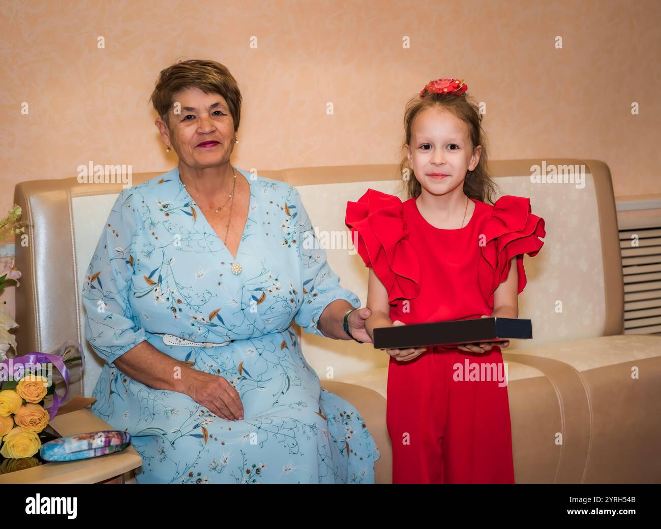 Eine 8-jährige Enkelin feiert ihren Geburtstag in einem roten Outfit mit einer Schleife. Ihre Großmutter, in festlichem Kleid gekleidet, gab ihr ein Geschenk. Das Mädchen Stockfoto