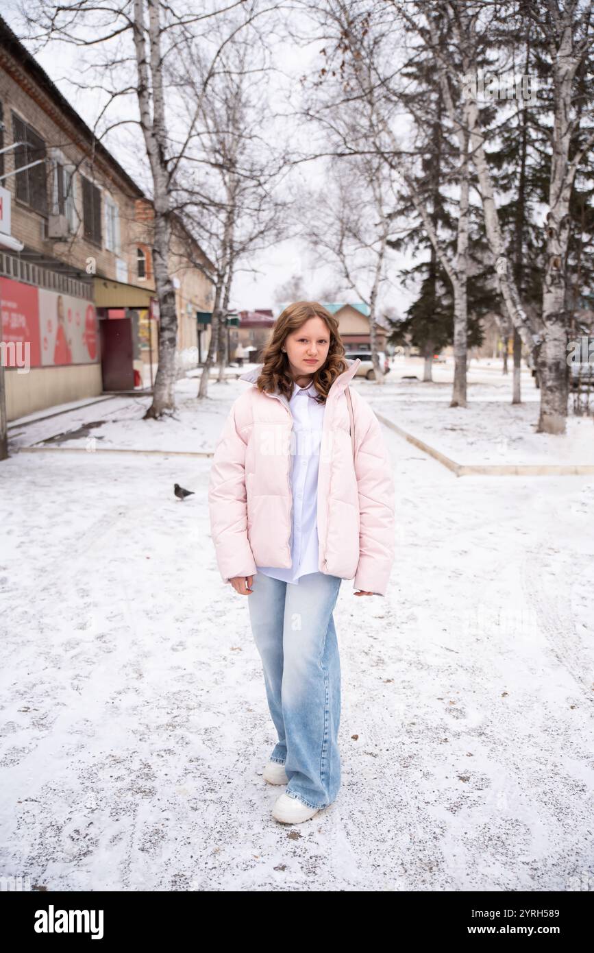 Das 15-jährige russische Mädchen in hellblauer Jeans und einer Winterjacke steht vor schneebedeckten Birken vor der Kulisse der Stadt. Sie strahlt Natur aus Stockfoto