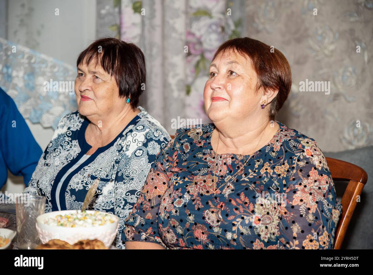 Zwei russische Frauen, eine 60 und die andere 70, schauen zur Seite auf einen Fernseher (auf dem Foto nicht zu sehen). Sie sind in festlicher Kleidung gekleidet, mit einer Ho Stockfoto