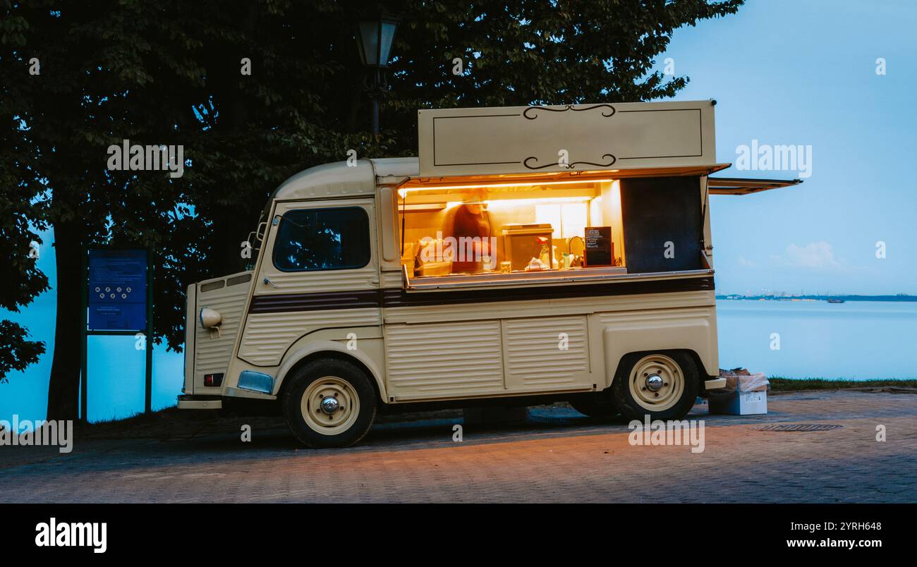 Vintage Food Truck parkt in der Nähe des Sees in der Dämmerung mit Koch, der im Inneren Speisen zubereitet, Street Food Business und Outdoor Catering Konzept Stockfoto