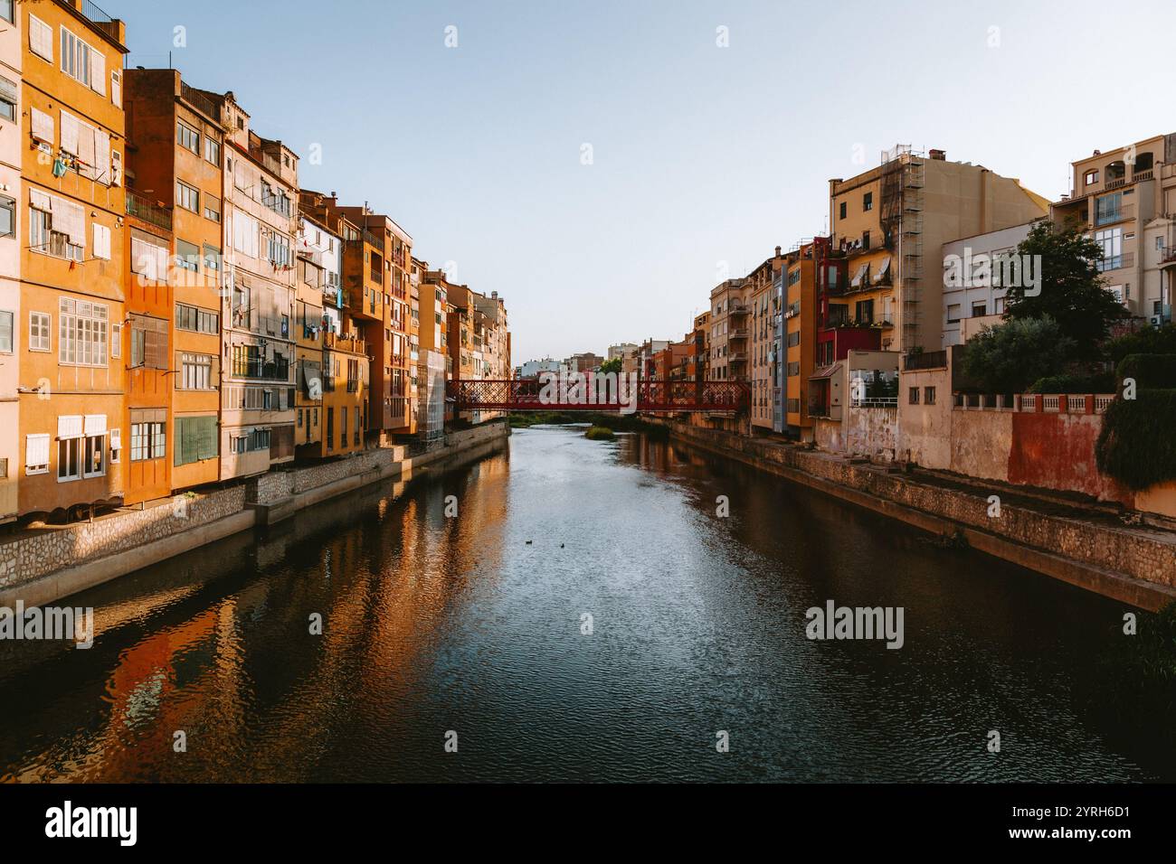 Der Fluss Onyar fließt friedlich unter der roten Brücke und reflektiert bei Sonnenuntergang warme Farbtöne traditioneller Gebäude an den Ufern in girona, katalonien, Spanien Stockfoto