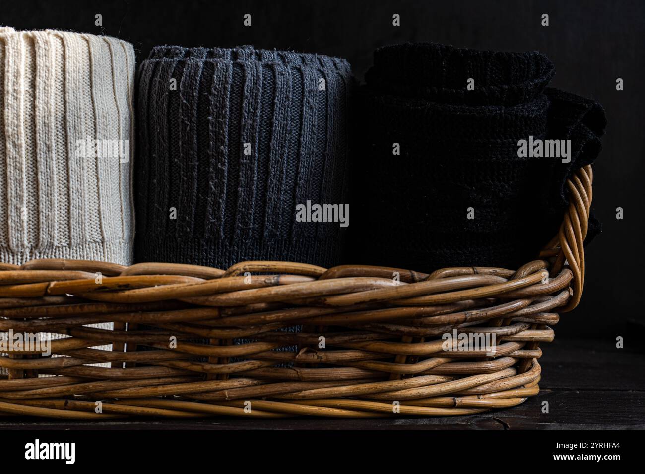 Weiche, strukturierte Strickstoffe in Schwarz, Grau und Creme sind ordentlich gerollt und in einem rustikalen Korb vor dunklem Holzhintergrund ausgestellt Stockfoto