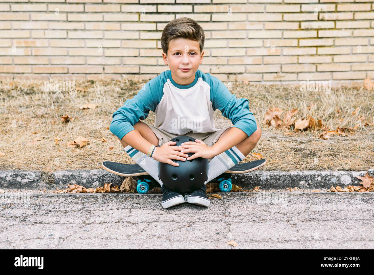 Ein kleiner Junge sitzt mit Skateboard und Helm auf dem Bürgersteig und trägt ein lässiges T-Shirt und Shorts. Der Hintergrund ist eine Ziegelwand und ein trockener Leaf Stockfoto