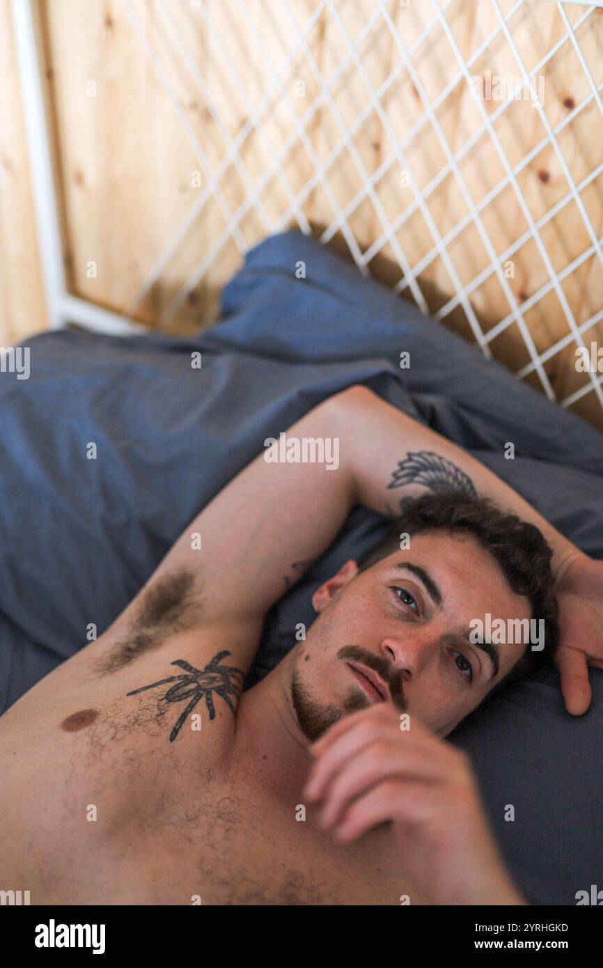 Ein Mann mit komplizierten Tätowierungen liegt bequem auf einem Bett mit blauen Laken, umgeben von Holzwänden und einem weißen Gitter. Die Szene vermittelt Entspannung Stockfoto