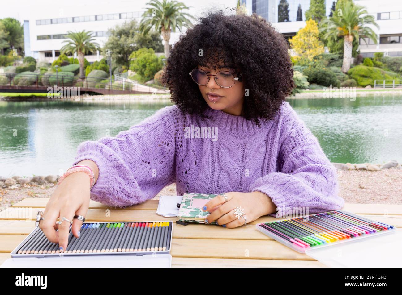 Ein Afro-Latino-Mädchen in einem lila Pullover ist mit leuchtenden Bleistiften gefärbt und genießt einen ruhigen Moment an einem malerischen See, umgeben von üppigem Grün Stockfoto