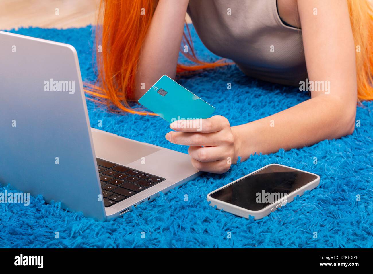 Eine Frau mit hellen Friseurgeschäften im Internet, mit einer türkisfarbenen Kreditkarte, während sie einen Laptop benutzt Sie liegt auf einem leuchtend blauen Teppich, mit einem Smartphone nea Stockfoto
