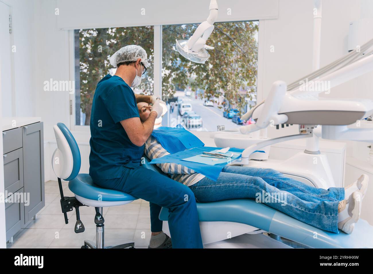 Ein Zahnarzt in blauen Peelings führt einen zahnärztlichen Eingriff an einem Patienten in einer gut ausgestatteten Klinik durch. Das helle Zimmer und der Blick auf die Straße schaffen einen professionellen geldautomaten Stockfoto