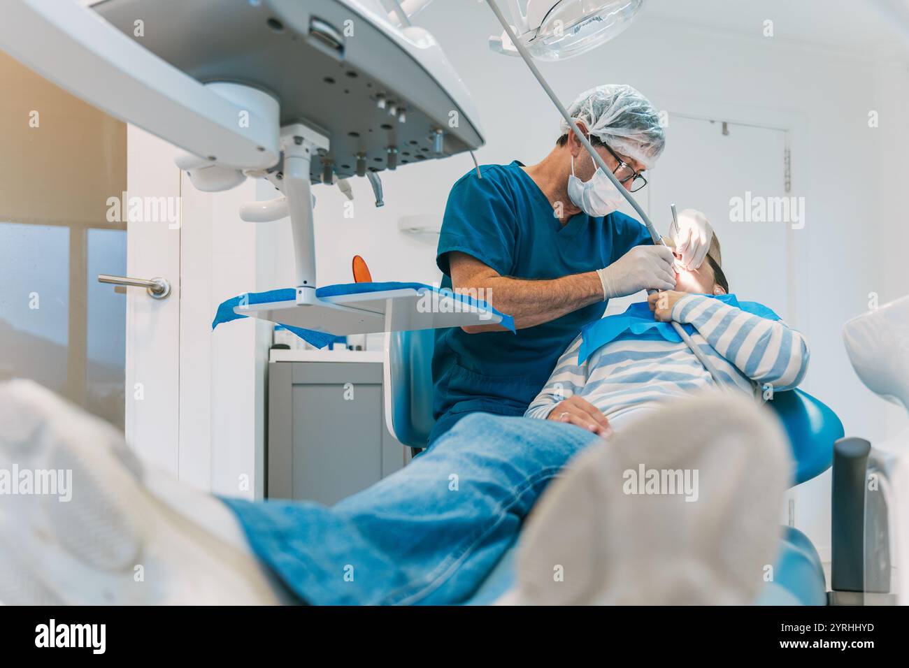 Ein Zahnarzt in blauen Peelings und einer chirurgischen Maske führt an einem Patienten in einer modernen Klinik zahnärztliche Arbeit durch. Der Patient lehnt sich in einem Zahnarztstuhl unter br Stockfoto